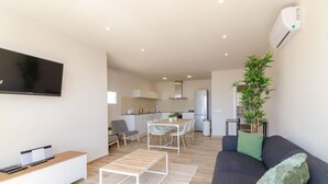 A modern living room with a grey sofa, a coffee table, and a flat-screen TV. There is a dining area with a table and chairs, and a kitchen in the background. - Club Villamar - (Lloret de Mar)