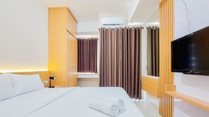 A hotel room with a neatly made bed, a bedside lamp, a flat-screen TV mounted on the wall, and a window with curtains. - Minimalist Studio Apartment At Serpong Garden (Cisauk)