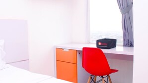 Habitación de hotel con una cama blanca, un escritorio naranja, una silla roja y una ventana con vista a la ciudad.