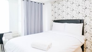 A neatly made bed with white linens and a rolled towel, a black headboard, and a window with a light blue curtain. - Clean Studio With Connected To Mall At Orchard Supermall Mansion Apartment (Surabaya)