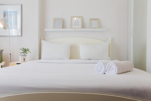 A neatly made bed with white linens and two rolled towels, a bedside table with a plant, and a wall-mounted shelf with three framed pictures. - Nice And Minimalist Studio Casa De Parco Apartment Bsd (Pagedangan)
