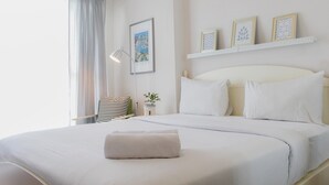 Un lit bien fait, avec du linge de lit blanc et une serviette pliée, une lampe de chevet, une petite plante en pot et un tableau encadré au mur.