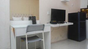 Una habitación compacta con un escritorio de vidrio, una silla gris, un televisor negro y un refrigerador negro.