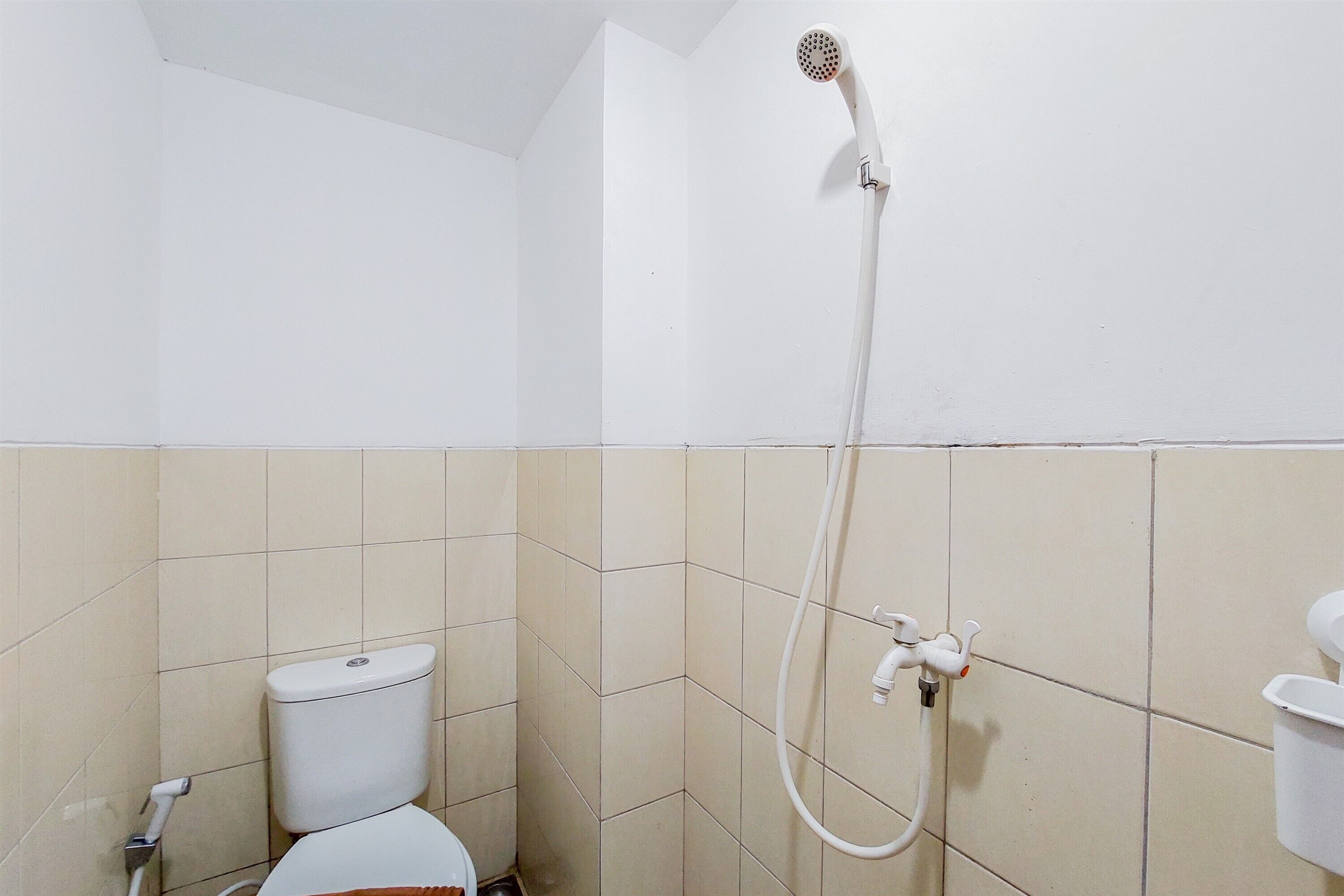 A small, white-walled bathroom with a toilet, a showerhead, and a wall-mounted faucet.