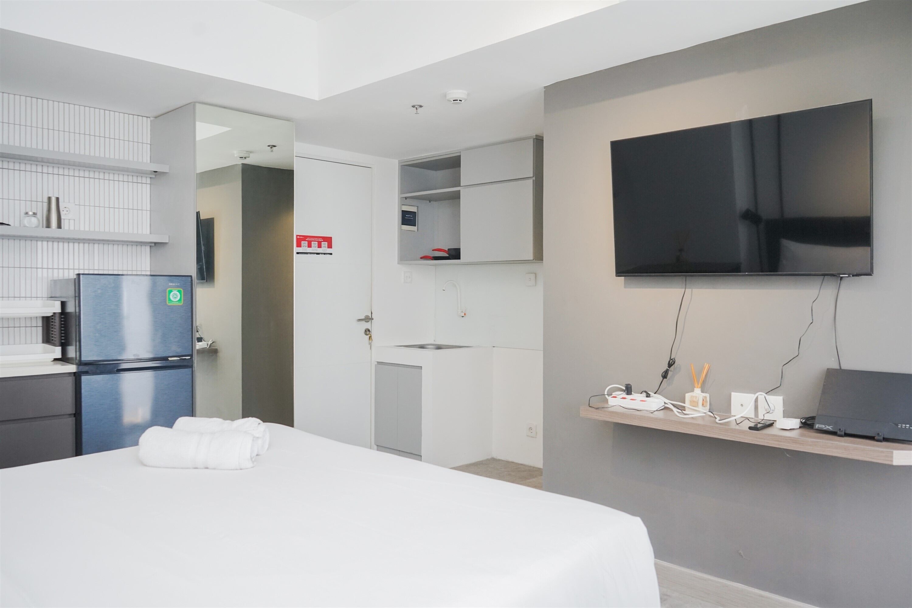 A modern hotel room with a bed, a refrigerator, a TV, a desk with a laptop, and a small kitchen area.