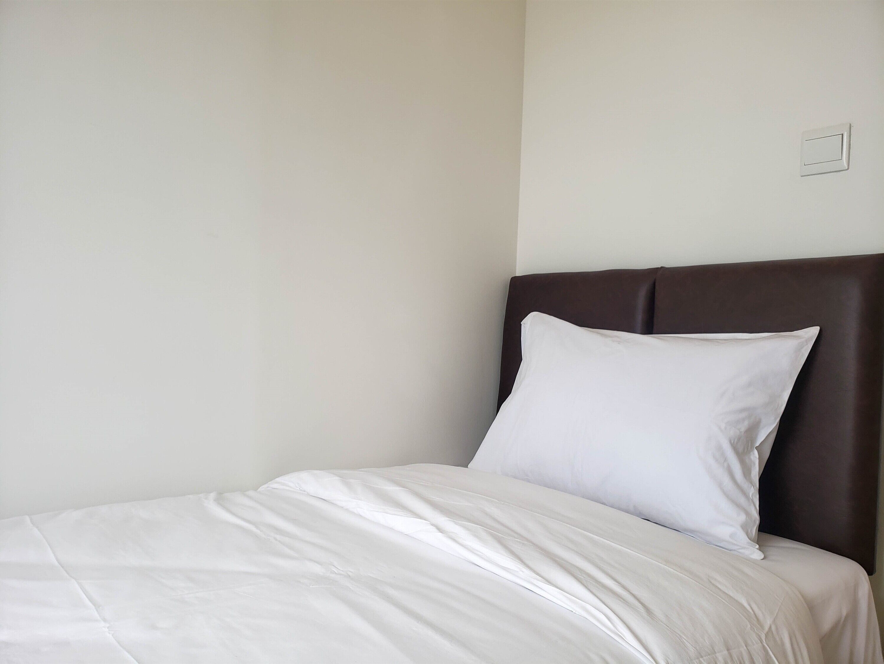 A neatly made bed with white bedding and a dark brown headboard in a simple room.