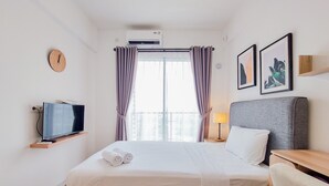 A hotel room with a bed, a TV on a shelf, a clock, air conditioning, curtains, and two framed pictures on the wall. - Warm And Simply Studio Apartment At Sky House Bsd (Pagedangan)