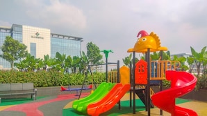 A playground with slides and a climbing structure, surrounded by greenery and a building in the background. - Nice And Homey Studio At Sayana Bekasi Apartment (Bekasi)