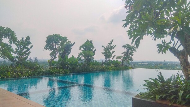 A swimming pool with a tiled edge, surrounded by lush greenery and trees. - Nice And Homey Studio At Sayana Bekasi Apartment (Bekasi)