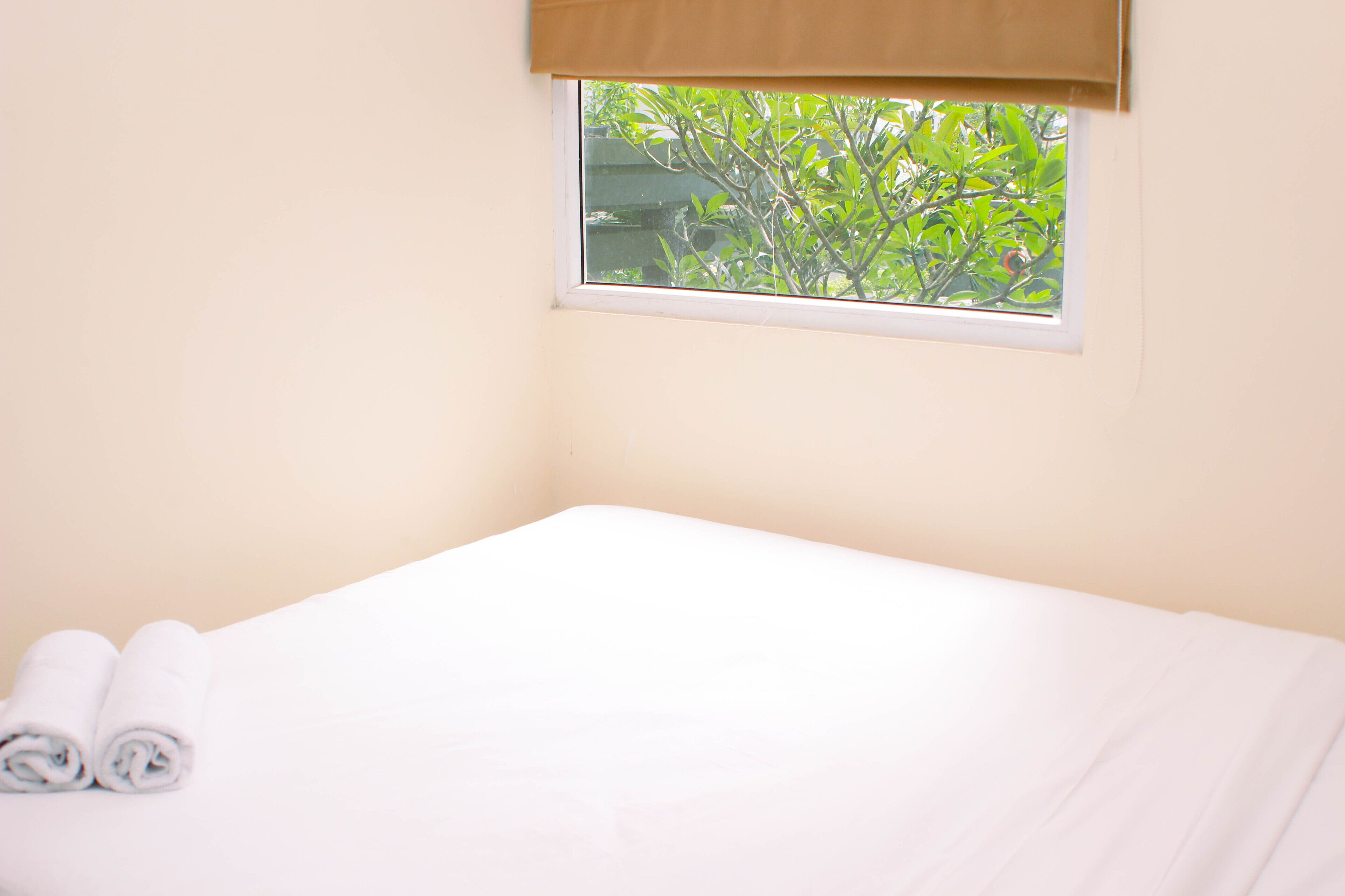 A clean, unoccupied room with a single bed, a window with a view of greenery, and a rolled towel on the bed.