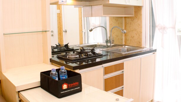 A compact kitchen with a gas stove, sink, and a black basket labeled 'travelio' on the counter. - Nice And Strategic 2Br At Green Pramuka City Apartment (Jakarta)