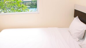 A neatly made bed with white linens and a wooden headboard, positioned next to a window with a view of greenery. - Nice And Strategic 2Br At Green Pramuka City Apartment (Jakarta)
