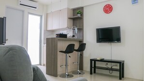 A modern living room with a grey sofa, a TV mounted on the wall, and a kitchen area with a wooden cabinet and two black bar stools. - Stunning And Comfy 2Br Bassura City Apartment (Jakarta)