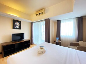 A hotel room with a large bed, a TV on a wooden stand, a small table with water bottles, and a chair. - Cozy 1Br Apartment At Braga City Walk (Bandung)