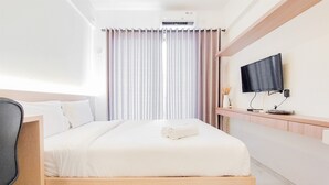A hotel room with a bed, a desk, a TV mounted on the wall, and a window with curtains. - Comfy Studio Room At 31St Floor Sky House Bsd Apartment (Pagedangan)