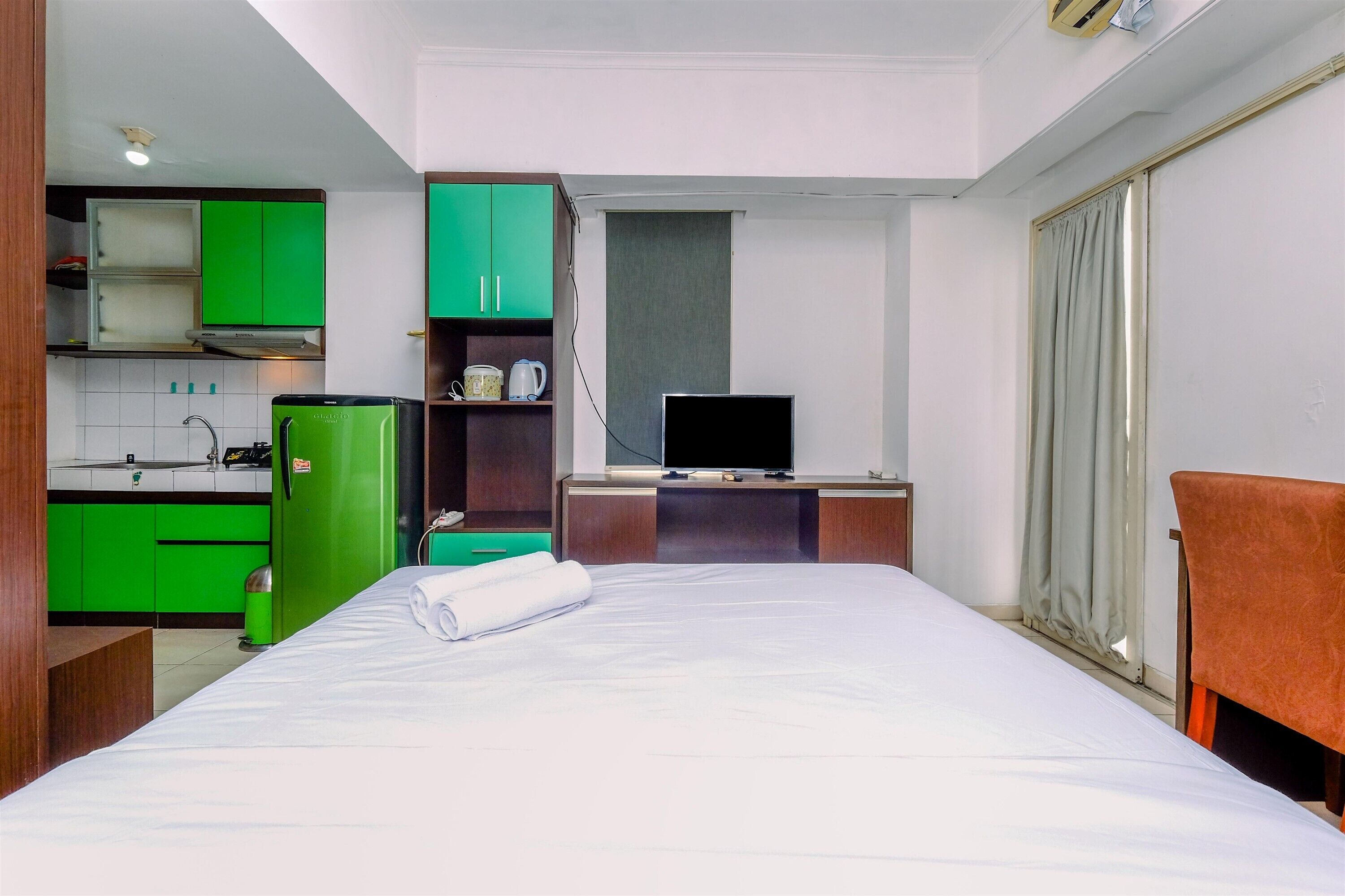 A compact hotel room with a bed, a desk with a television, a kitchenette, and a window with curtains.