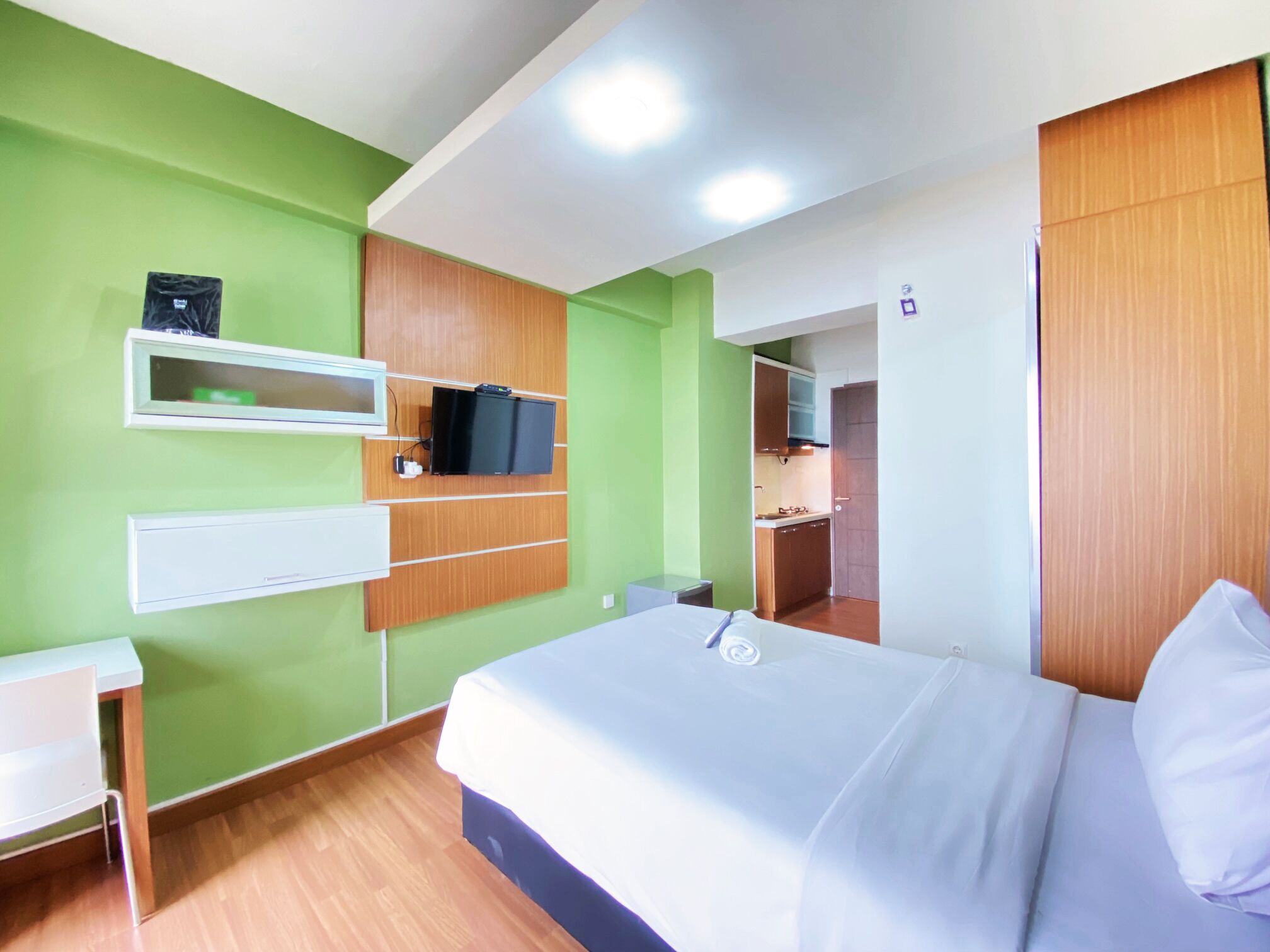 A hotel room with a bed, a desk, a TV mounted on the wall, and a kitchenette area.