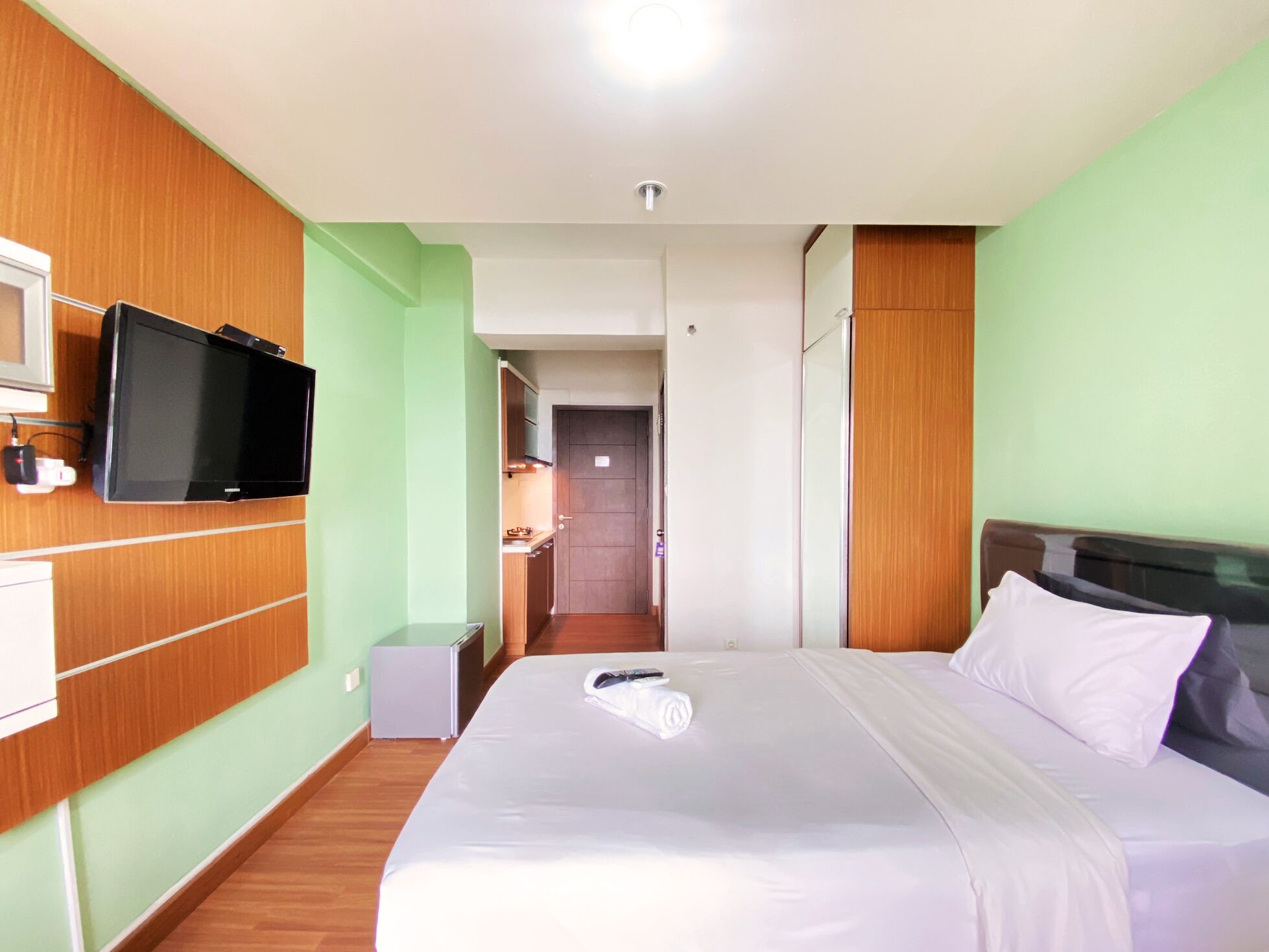 Ein Hotelzimmer mit einem Bett, einem an der Wand befestigten Fernseher, einer kleinen Küchenzeile und einer Tür zu einem weiteren Raum.