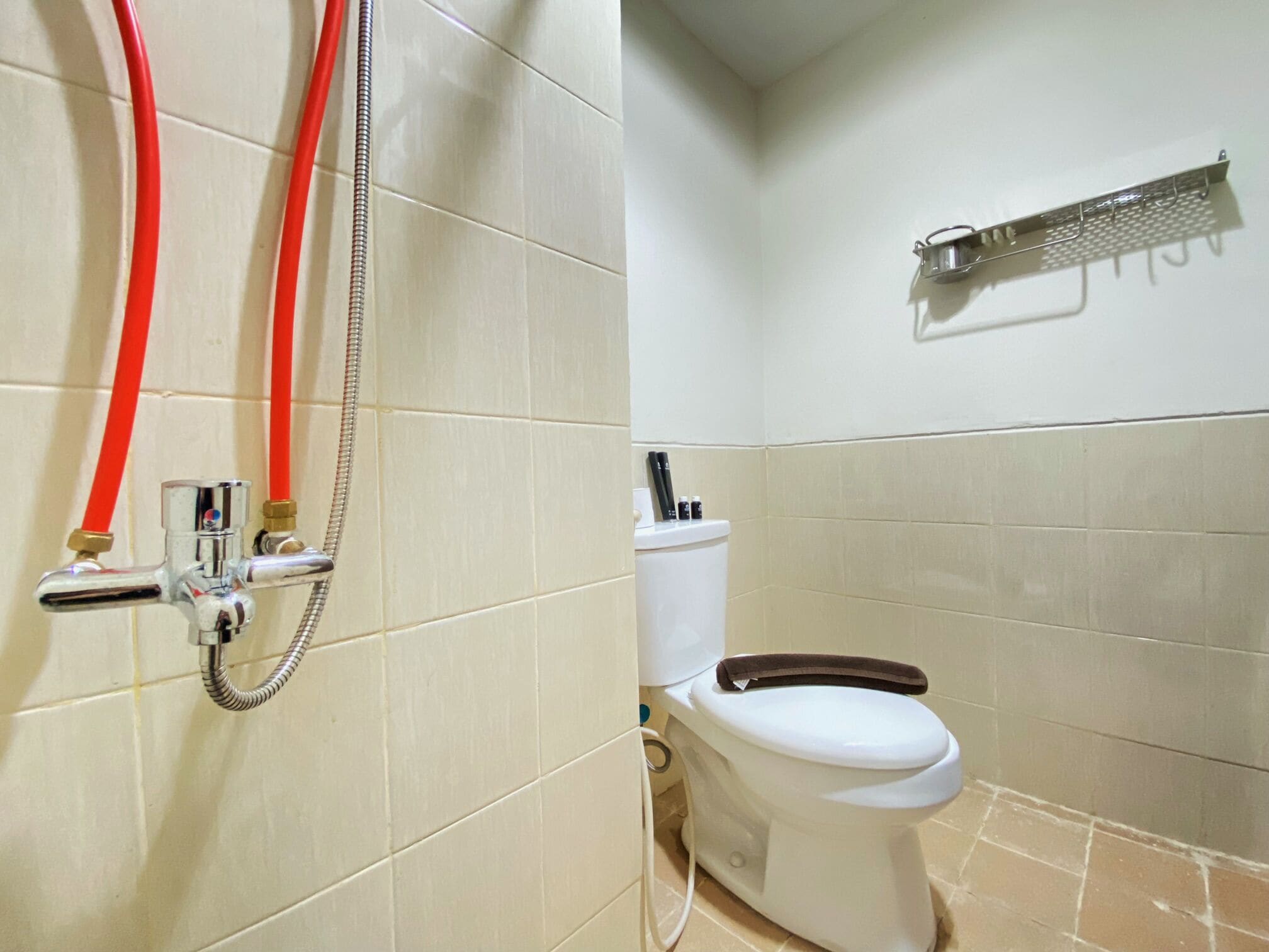 A bathroom with a toilet, a shower with red hoses, and a towel rack on the wall.
