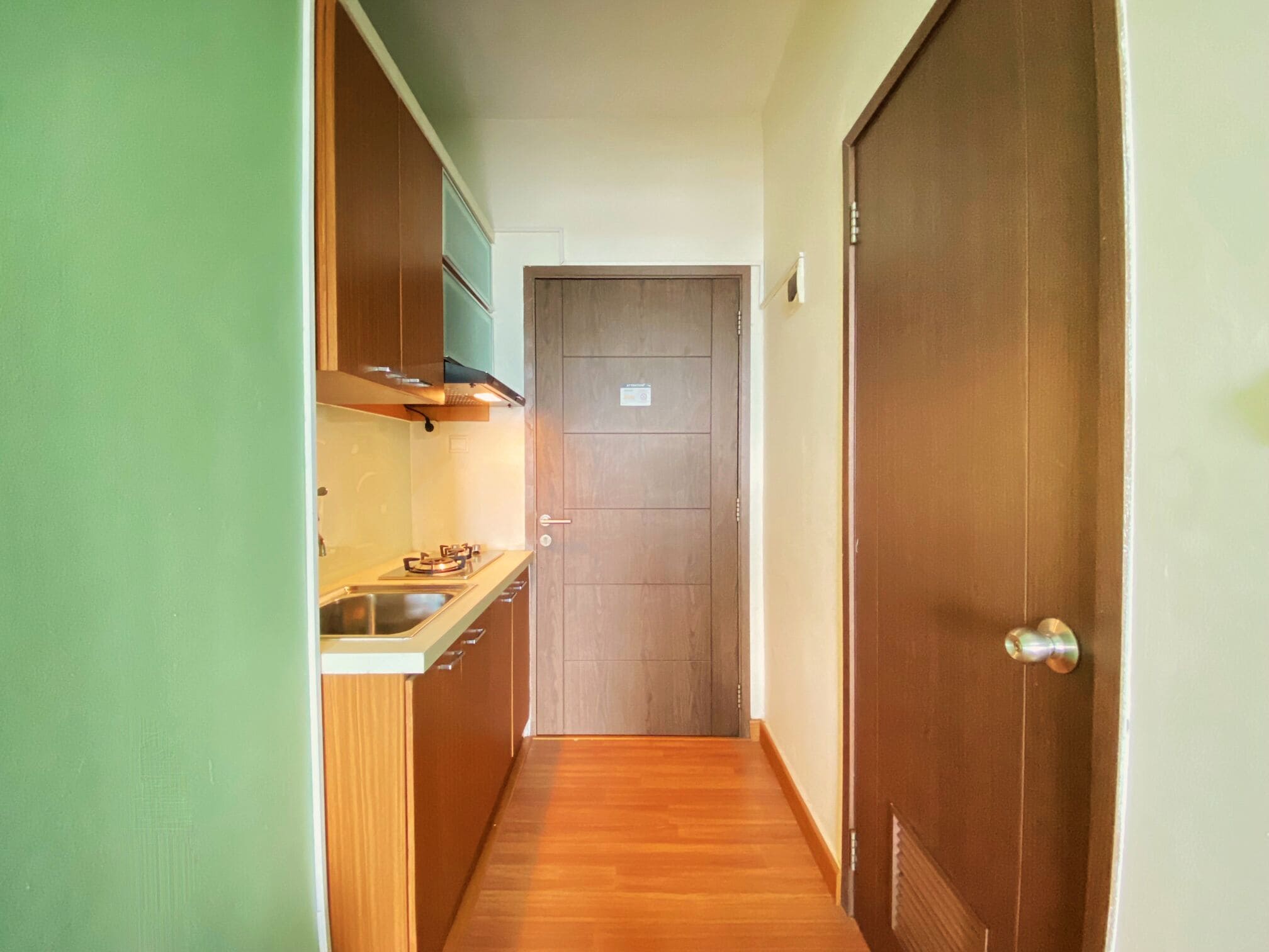 A narrow hallway with a wooden floor, a kitchen area with a stove and cabinets, and a closed door at the end.