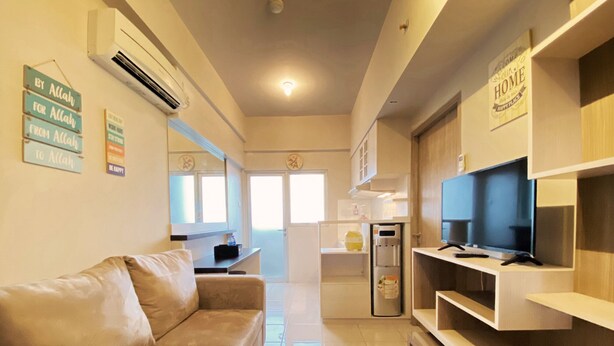 A modern living room with a beige sofa, a flat-screen TV on a wooden stand, and a kitchen area in the background. - Relaxing 1Br At Apartment At Newton Residence Bandung (Bandung)