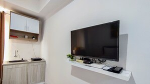 Una habitación moderna con un televisor de pantalla plana montado en la pared, una pequeña cocina con pileta y alacenas, y un estante blanco debajo del televisor que contiene dispositivos electrónicos.