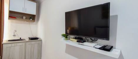 A modern room with a flat-screen TV mounted on the wall, a small kitchen area with a sink and cabinets, and a white shelf below the TV holding electronic devices.