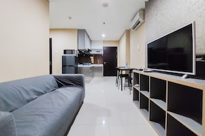 Uma sala de estar moderna com um sofá cinzento, uma TV sobre um suporte de madeira e uma cozinha ao fundo.