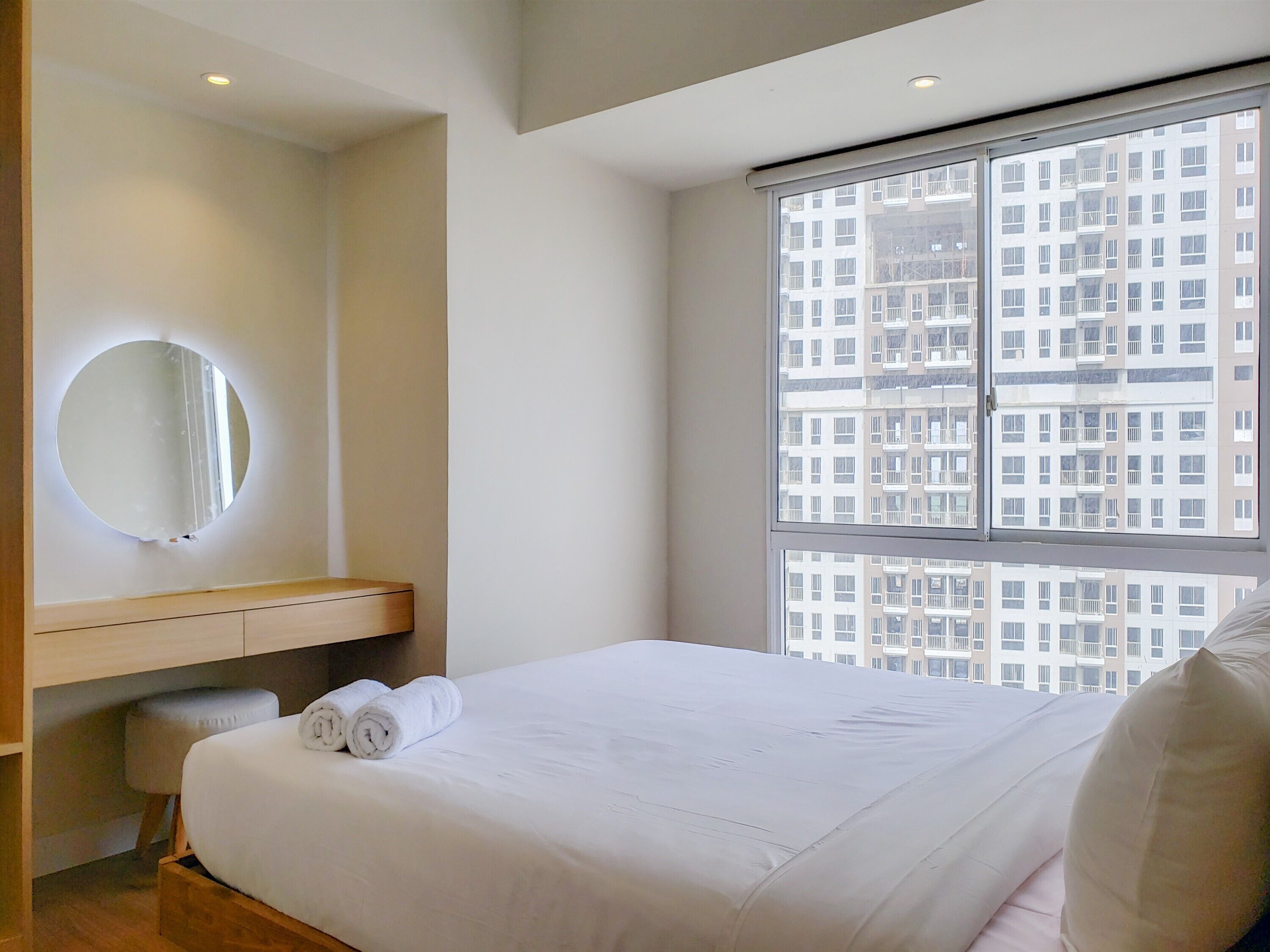 Ein modernes Hotelzimmer mit einem großen Bett, einem runden Spiegel, einer hölzernen Bank und Blick auf ein Hochhaus.