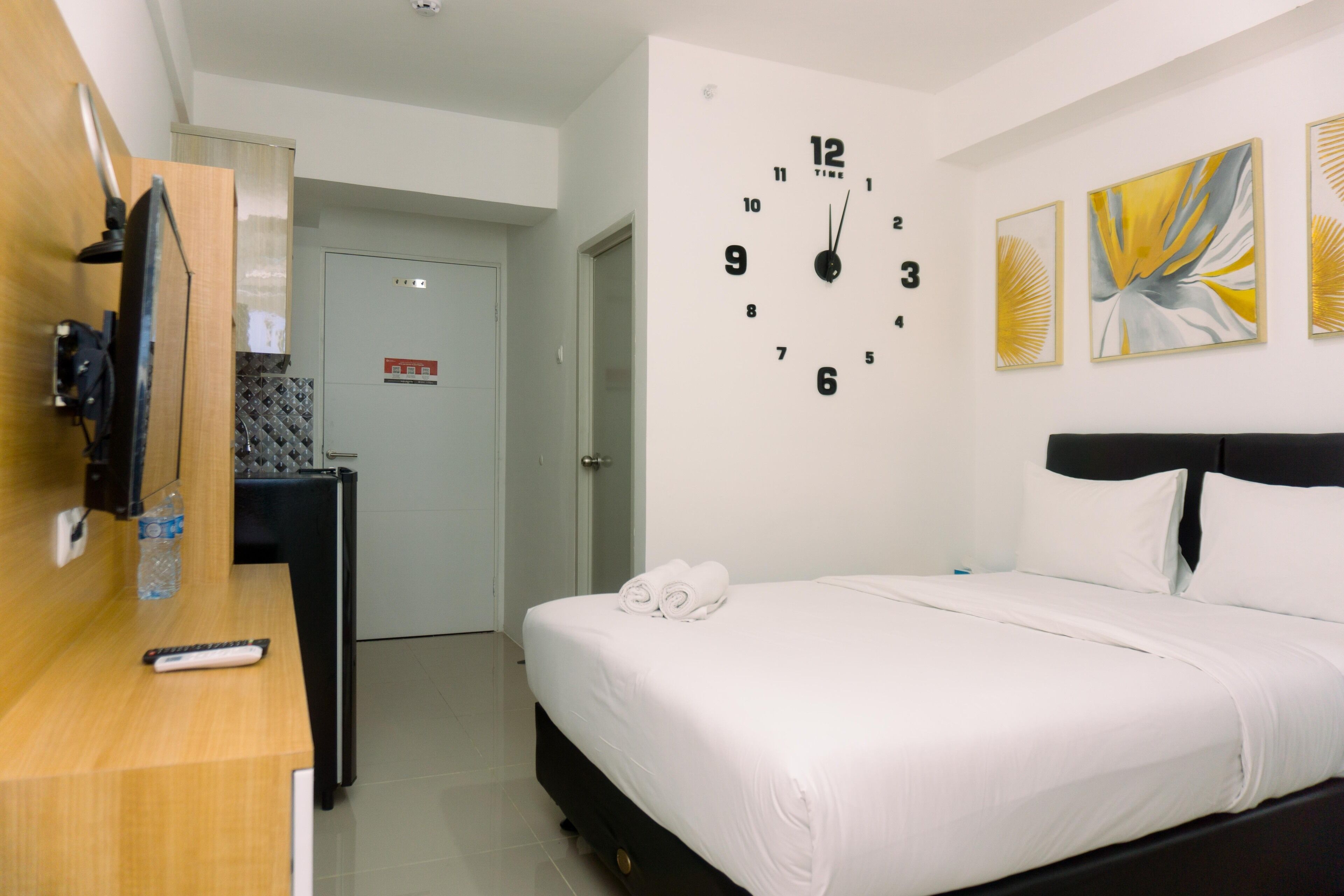 A hotel room with a bed, a wall clock, artwork, a desk with a remote control, and a door.