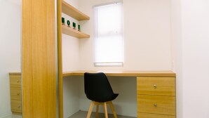 A corner bedroom with a wooden desk, a black chair, a small shelf with decorative items, and a window with blinds. - Cozy Studio At Urbantown Serpong Apartment (South Tangerang)