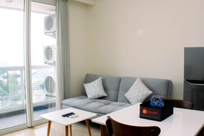 A modern hotel room with a grey sofa, a small white table, and a dining table. There are two air conditioning units on the wall, a black box with water bottles on the dining table, and a TV on the wall.
