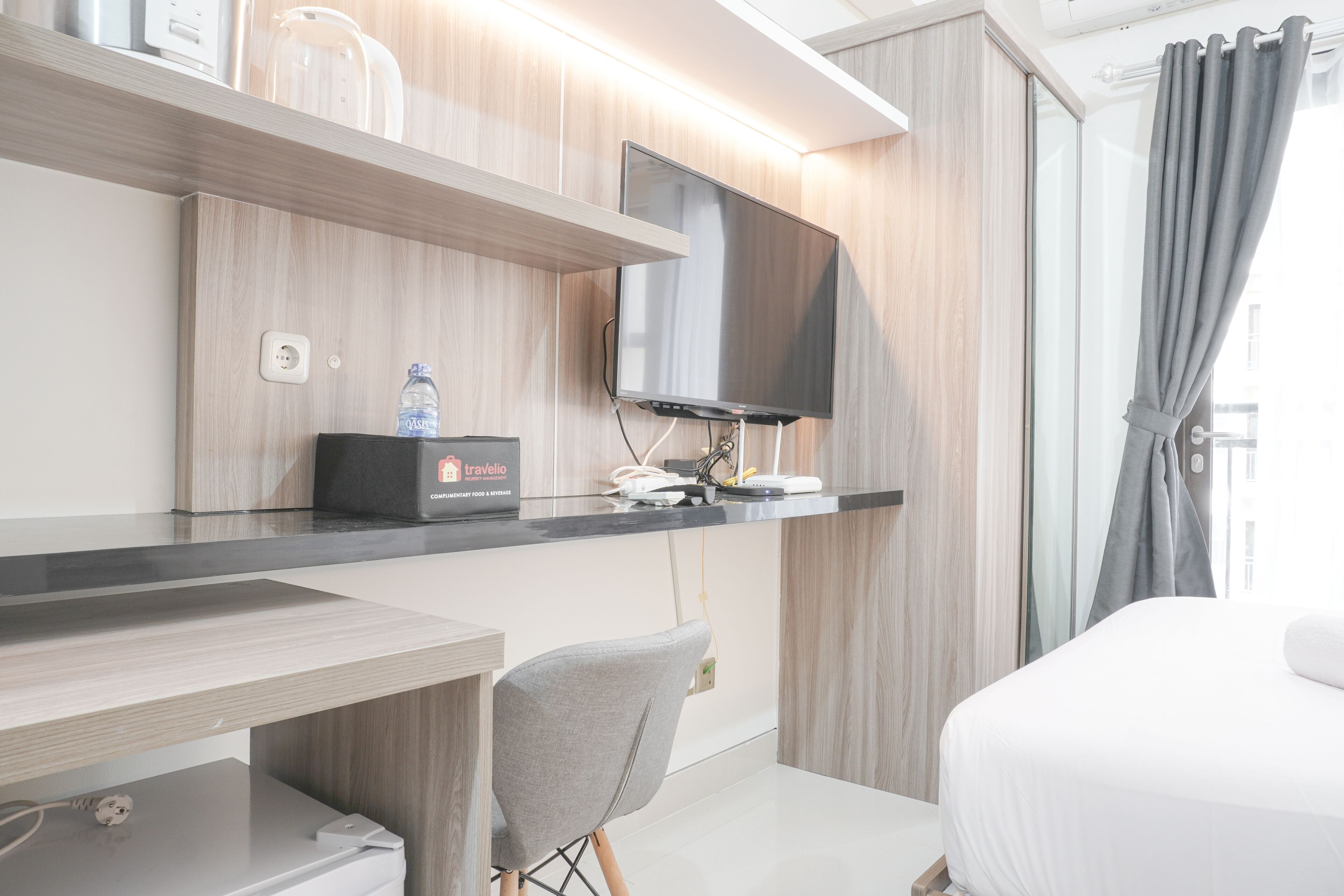 A modern hotel room with a flat-screen TV mounted on the wall, a desk with a chair, a bed, and a large window with curtains.