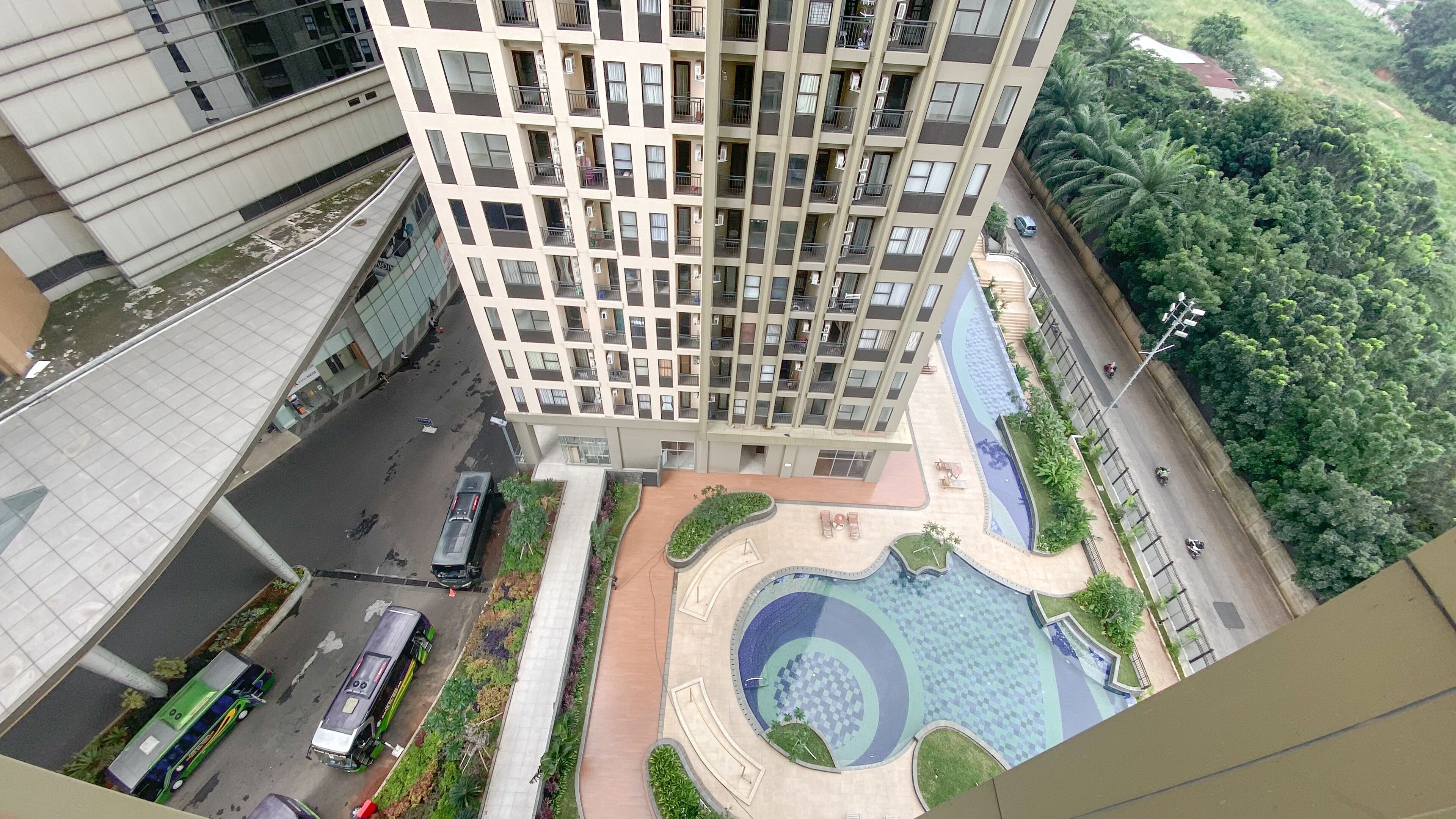 A high-rise building with multiple balconies, a landscaped courtyard with a pool, and a road with vehicles.