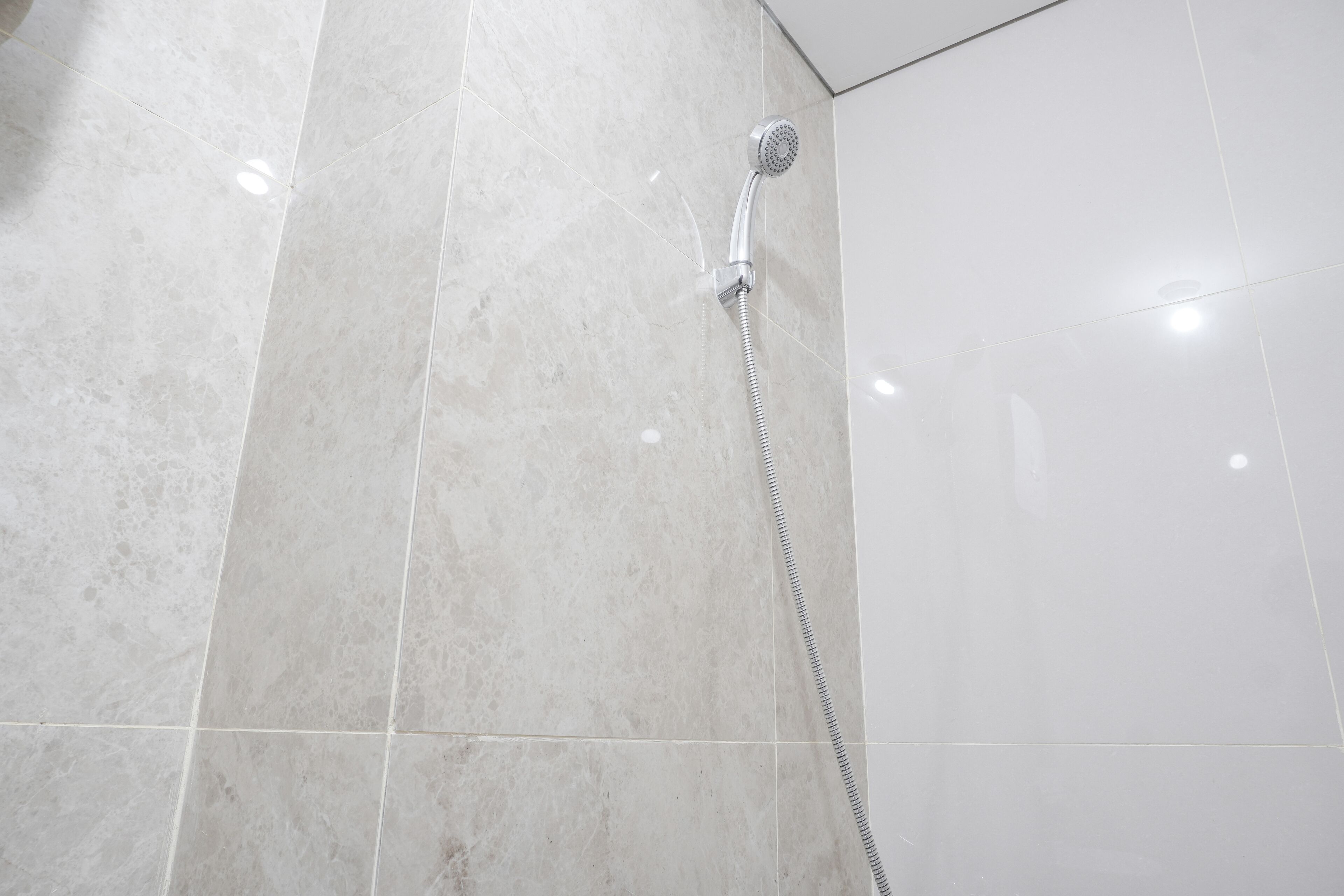 A bathroom with a shower area featuring a chrome shower head and a flexible hose. The walls are tiled with a light-colored marble.