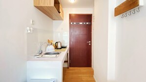 Cocina compacta con armarios blancos, un lavabo, una tetera y una cafetera. Hay una puerta de madera y un pequeño banco de madera.