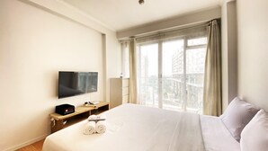 Una habitación de hotel con una cama grande, un televisor de pantalla plana en la pared, una mesita de noche con lámpara y una ventana con cortinas.