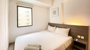 A hotel room with a bed, a bedside table with a phone, a window with blinds, and a framed picture on the wall. - Fancy Designed 2Br At Gateway Ahmad Yani Cicadas Apartment (Bandung)