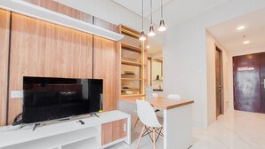 A modern living room with a flat-screen TV, wooden cabinets, a white entertainment unit, and a small dining area with a white chair. - Well Designed And Comfy 2Br Sky House Bsd Apartment (Pagedangan)