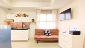 A compact kitchen with white cabinets, a refrigerator, and a tiled backsplash. There is a sofa with a cushion and a flat-screen TV mounted on the wall. - Comfy And Modern 2Br Apartment At Parahyangan Residence (Bandung)