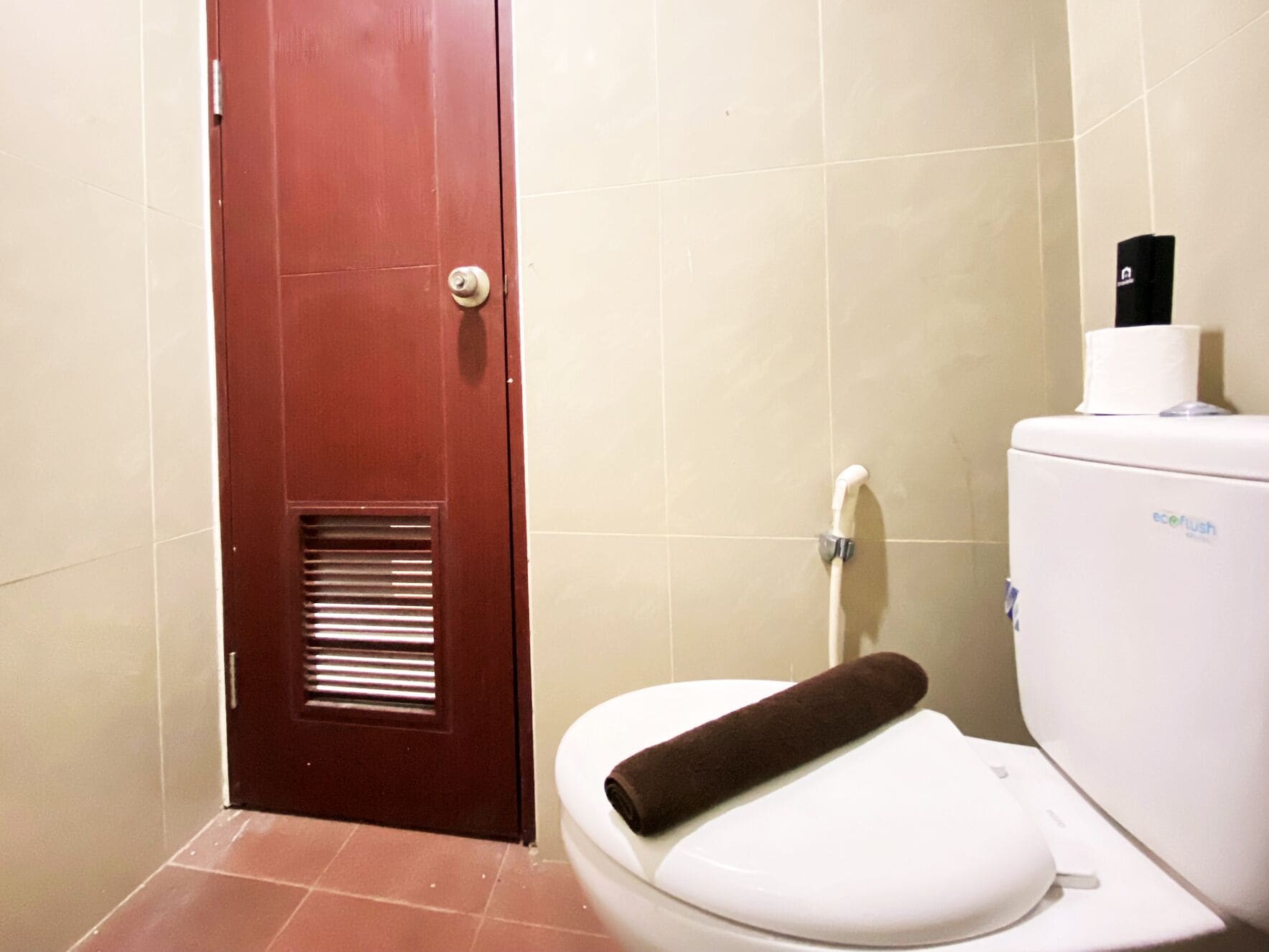 Un bany amb un vàter blanc, una porta marró i un suport de paper higiènic blanc.