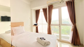 A hotel room with a bed, a TV, a window with curtains, and an air conditioning unit. - Warm And Cozy Studio At Taman Melati Jatinangor Apartment (Hegarmanah)