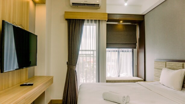 A hotel room with a bed, a TV on a wooden console, an air conditioner, and a window with curtains. - Minimalist And Warm Studio At Transpark Bintaro Apartment (South Tangerang)