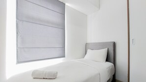 A modern bedroom with a bed, a grey headboard, and a window with blinds. - Luxurious 2Br At Serpong Garden Apartment (Cisauk)