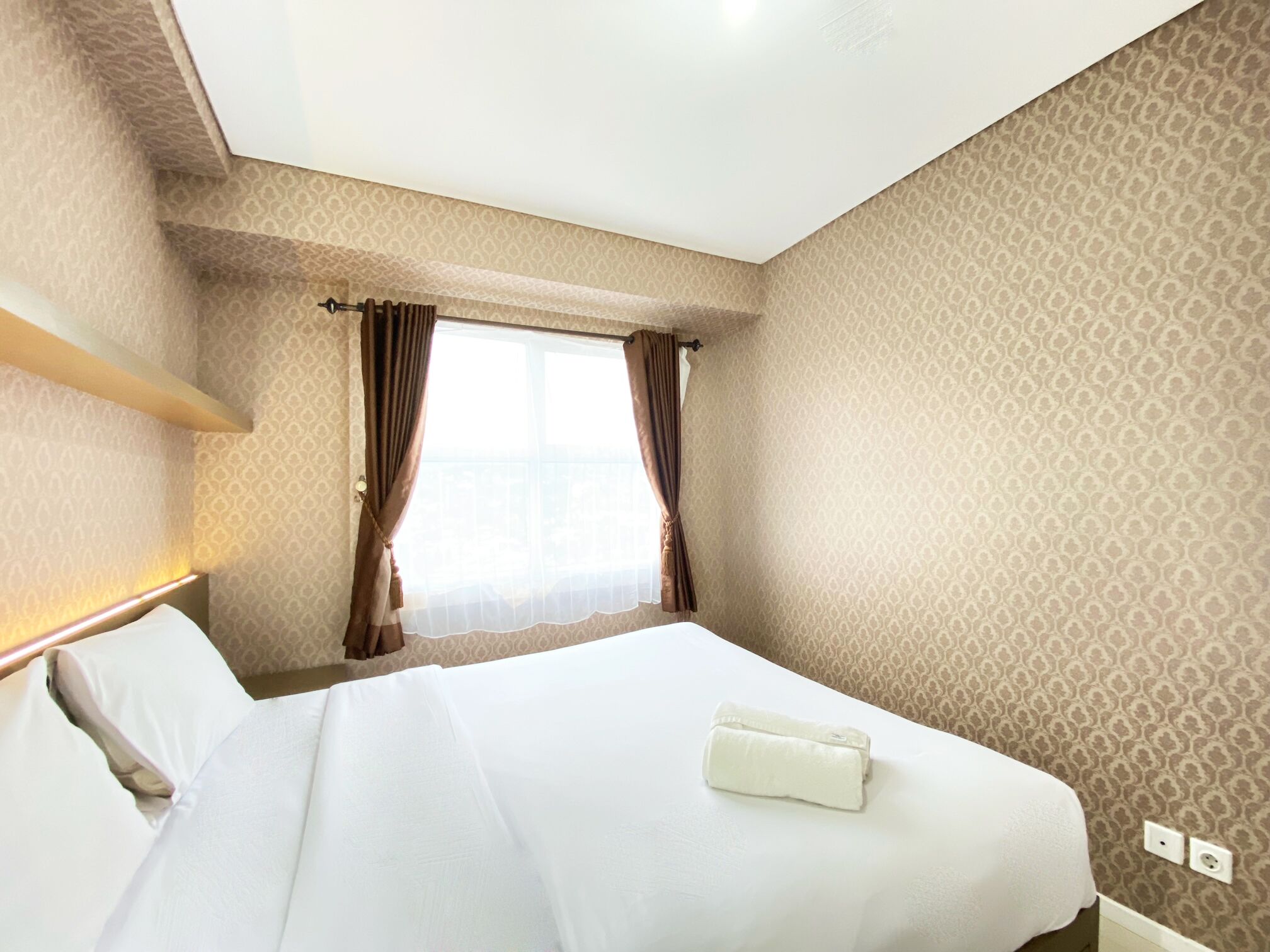 A hotel room with a neatly made bed, a window with curtains, and patterned wallpaper.