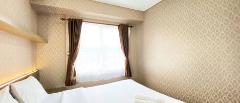 A hotel room with a neatly made bed, a window with curtains, and patterned wallpaper.