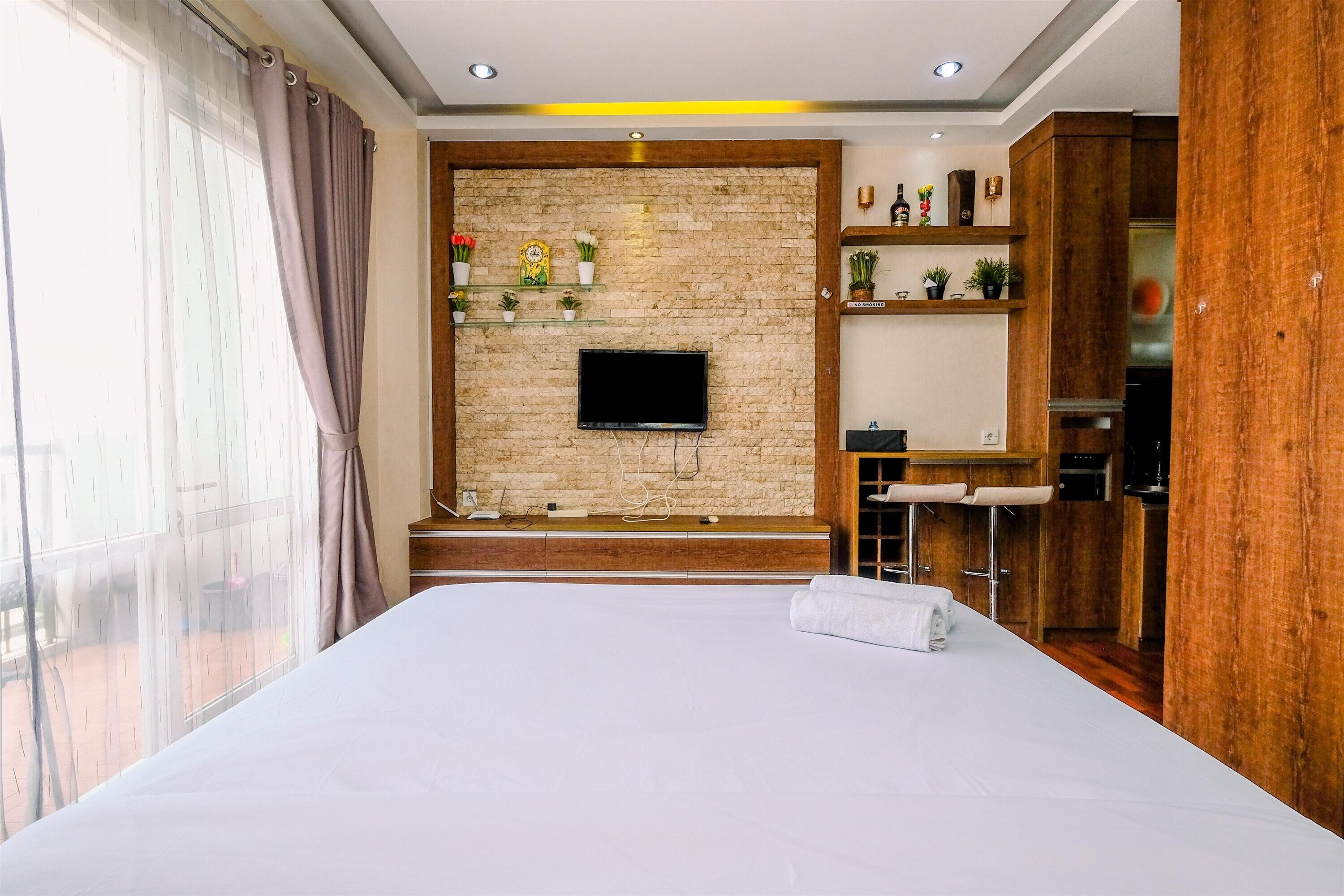 A hotel room with a large bed, a TV mounted on a brick wall, a small kitchen area, and a balcony with curtains.