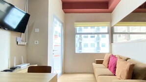 Una habitación con un sofá color beige, un televisor en la pared, una mesa de madera y una ventana con vistas a edificios.