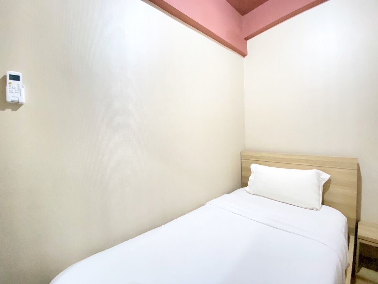 Un lit simple avec une tête de lit en bois, dans une pièce aux murs unis et dont le plafond est orné d’une poutre rose.