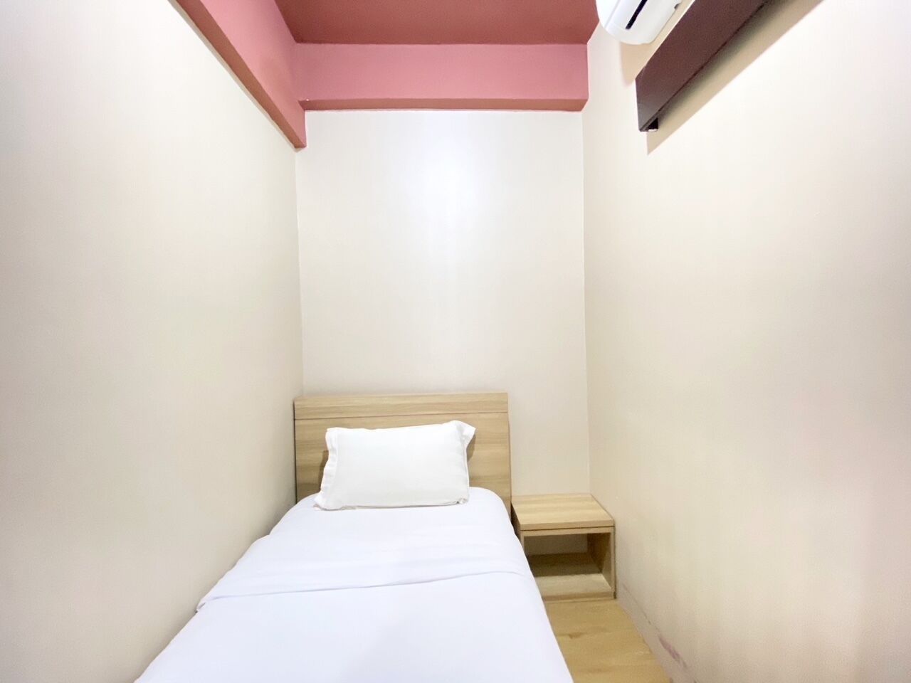 Une petite pièce étroite, avec un lit simple, une table de chevet en bois et un climatiseur fixé au mur.